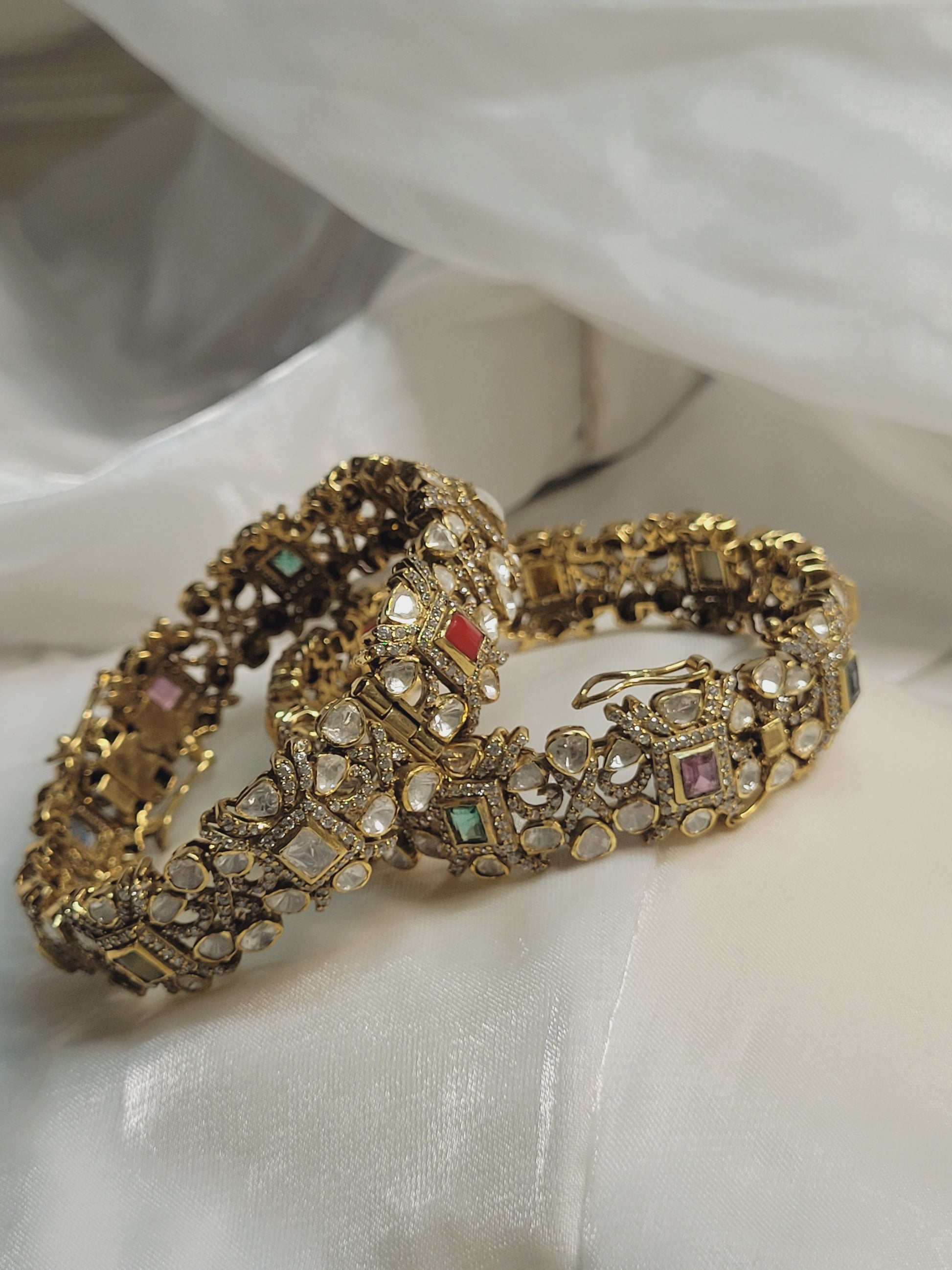 Pair of ornate gold bangles embellished with multicolored gemstones, resting elegantly on soft white draped fabric.