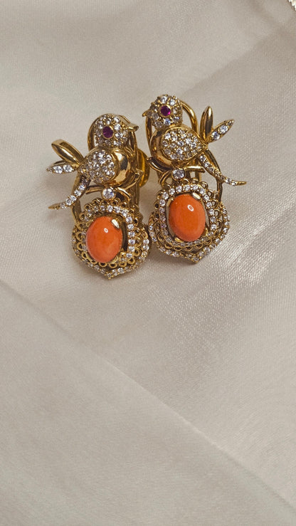 Gold bird-shaped earrings with orange stones and diamond accents on a satin surface