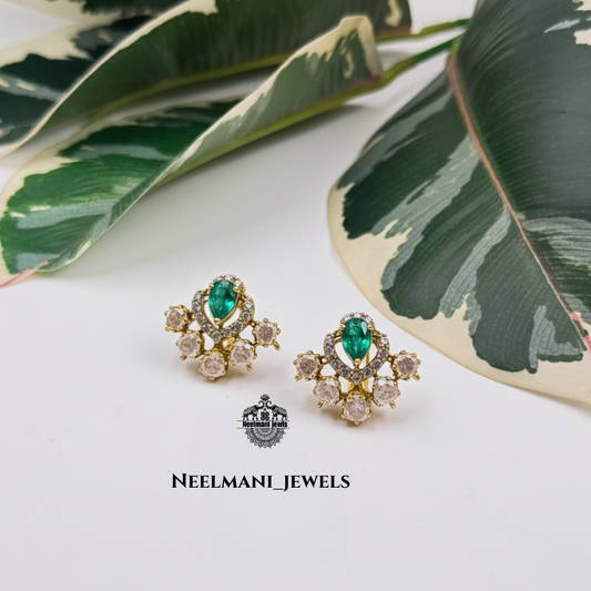 Gold earrings with green gemstones on a white surface with leaves in the background, featuring Neelmani Jewels branding.