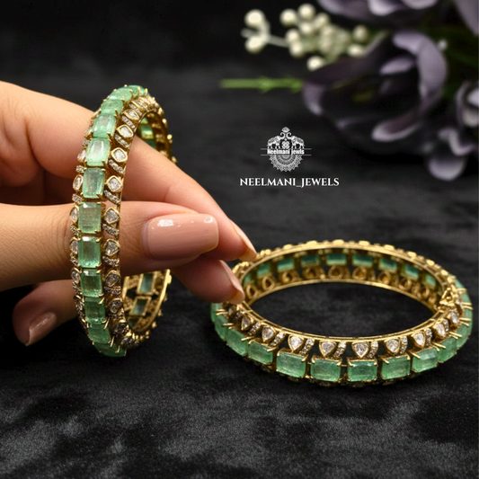 Gold and green emerald bangles held by a hand on a dark background with Neelmani Jewels logo.